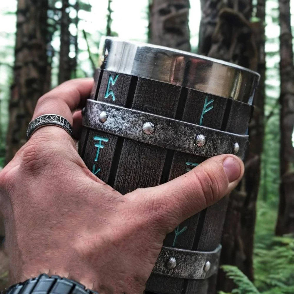 Hand holding wooden mug with metal bands and runes in forest.